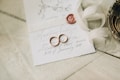 A close-up of an elegantly arranged wedding invitation featuring handwritten calligraphy. Two gold wedding rings are placed on the card alongside a red wax seal. A cream-colored ribbon is artistically draped nearby, and a decorative metal dish is partially visible.