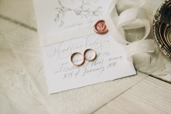 A close-up of an elegantly arranged wedding invitation featuring handwritten calligraphy. Two gold wedding rings are placed on the card alongside a red wax seal. A cream-colored ribbon is artistically draped nearby, and a decorative metal dish is partially visible.