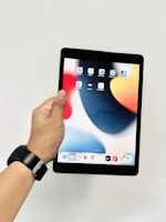 A hand holding a black tablet with a colorful home screen displaying various app icons. The person is wearing a black smartwatch. The background is plain and white.
