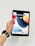 A hand holding a black tablet with a colorful home screen displaying various app icons. The person is wearing a black smartwatch. The background is plain and white.