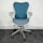 A modern, ergonomic office chair with a blue mesh backrest and seat. It features light gray armrests and base, with five wheels for mobility. The chair is positioned on dark gray carpet against a plain, white wall.