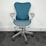 A modern, ergonomic office chair with a blue mesh backrest and seat. It features light gray armrests and base, with five wheels for mobility. The chair is positioned on dark gray carpet against a plain, white wall.