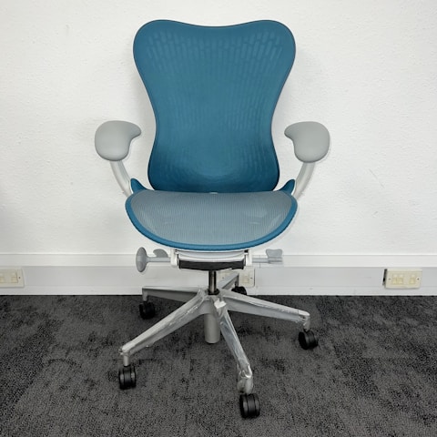 A modern, ergonomic office chair with a blue mesh backrest and seat. It features light gray armrests and base, with five wheels for mobility. The chair is positioned on dark gray carpet against a plain, white wall.