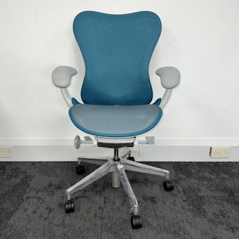 A modern, ergonomic office chair with a blue mesh backrest and seat. It features light gray armrests and base, with five wheels for mobility. The chair is positioned on dark gray carpet against a plain, white wall.