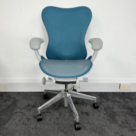 A modern, ergonomic office chair with a blue mesh backrest and seat. It features light gray armrests and base, with five wheels for mobility. The chair is positioned on dark gray carpet against a plain, white wall.