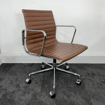 A modern office chair with a sleek design, featuring a brown padded seat and backrest made of leather or a similar material. The chair has metal armrests and a chrome-finished, five-point base with caster wheels, resting on a dark carpeted floor against a plain white wall.