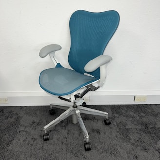 A modern ergonomic office chair with a blue mesh backrest and seat, featuring white armrests and a silver base with five rolling casters. It is positioned on a dark carpet in front of a white wall, near some electrical outlets.