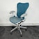 A modern ergonomic office chair with a blue mesh backrest and seat, featuring white armrests and a silver base with five rolling casters. It is positioned on a dark carpet in front of a white wall, near some electrical outlets.