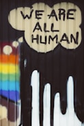 Graffiti art featuring the words 'We Are All Human' painted in bold, black letters inside a cloud-like shape on a corrugated surface. A rainbow with vibrant colors is displayed on the left side, suggesting themes of diversity and inclusivity.