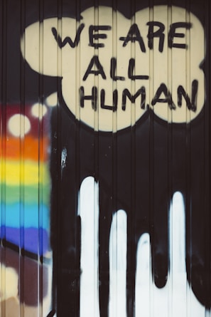 Graffiti art featuring the words 'We Are All Human' painted in bold, black letters inside a cloud-like shape on a corrugated surface. A rainbow with vibrant colors is displayed on the left side, suggesting themes of diversity and inclusivity.