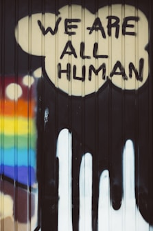 Graffiti art featuring the words 'We Are All Human' painted in bold, black letters inside a cloud-like shape on a corrugated surface. A rainbow with vibrant colors is displayed on the left side, suggesting themes of diversity and inclusivity.