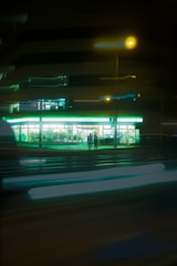 A convenience store with bright, glowing lights stands out against the night backdrop. Two people are visible in silhouette near the entrance, and light trails from passing vehicles blur in front, adding a dynamic element to the scene.
