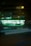 A convenience store with bright, glowing lights stands out against the night backdrop. Two people are visible in silhouette near the entrance, and light trails from passing vehicles blur in front, adding a dynamic element to the scene.
