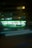 A convenience store with bright, glowing lights stands out against the night backdrop. Two people are visible in silhouette near the entrance, and light trails from passing vehicles blur in front, adding a dynamic element to the scene.