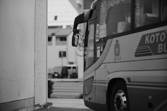 A bus is partially visible, parked close to a building with a tiled exterior. The bus is adorned with logos and text, including 'Koto' and 'President.' In the background, there are city buildings and a street with a vehicle parked nearby, all captured in black and white.