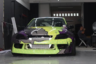 A car with a bright and colorful custom paint job is parked inside a garage. The car's front bumper and hood feature a geometric pattern with shades of lime green, black, and purple. A person is seated in the background on the right side of the image, next to a large fan and some equipment.