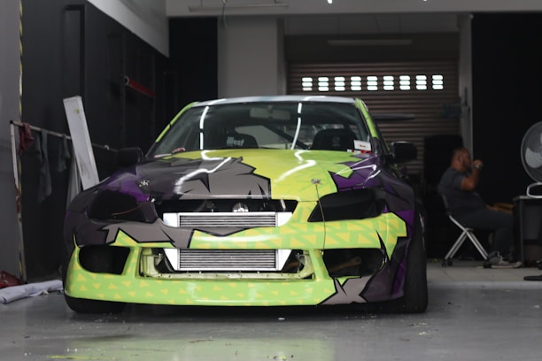A car with a bright and colorful custom paint job is parked inside a garage. The car's front bumper and hood feature a geometric pattern with shades of lime green, black, and purple. A person is seated in the background on the right side of the image, next to a large fan and some equipment.