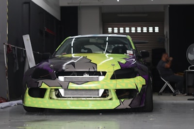 A car with a bright and colorful custom paint job is parked inside a garage. The car's front bumper and hood feature a geometric pattern with shades of lime green, black, and purple. A person is seated in the background on the right side of the image, next to a large fan and some equipment.