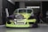 A car with a bright and colorful custom paint job is parked inside a garage. The car's front bumper and hood feature a geometric pattern with shades of lime green, black, and purple. A person is seated in the background on the right side of the image, next to a large fan and some equipment.