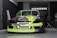 A car with a bright and colorful custom paint job is parked inside a garage. The car's front bumper and hood feature a geometric pattern with shades of lime green, black, and purple. A person is seated in the background on the right side of the image, next to a large fan and some equipment.