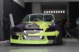 A car with a bright and colorful custom paint job is parked inside a garage. The car's front bumper and hood feature a geometric pattern with shades of lime green, black, and purple. A person is seated in the background on the right side of the image, next to a large fan and some equipment.