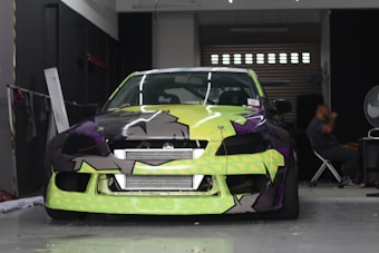A car with a bright and colorful custom paint job is parked inside a garage. The car's front bumper and hood feature a geometric pattern with shades of lime green, black, and purple. A person is seated in the background on the right side of the image, next to a large fan and some equipment.
