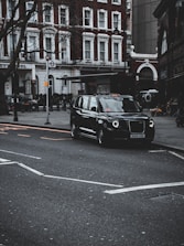 A classic black taxi is parked on a city street, with a backdrop of elegant red-brick buildings featuring white-trimmed windows. The street is lined with bare trees, and there's an iconic London Underground sign visible. The scene exudes an urban atmosphere, suggesting a typical day in a bustling city setting.