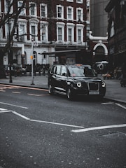 A sleek black taxi waiting outside a busy London airport terminal with a driver holding a welcome sign.