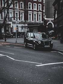 A classic black taxi is parked on a city street, with a backdrop of elegant red-brick buildings featuring white-trimmed windows. The street is lined with bare trees, and there's an iconic London Underground sign visible. The scene exudes an urban atmosphere, suggesting a typical day in a bustling city setting.