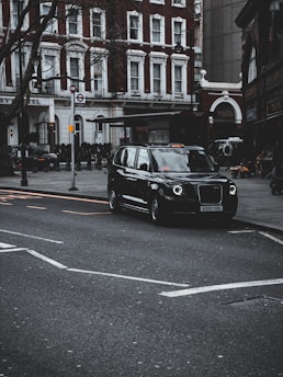 A sleek black taxi waiting outside a busy London airport terminal with a driver holding a welcome sign.