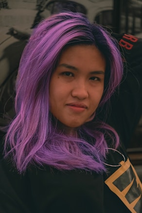 A person with long, vibrant purple hair is gazing at the camera with a subtle, confident expression. The hair has a smooth texture that adds a sense of vividness to the image. The individual is wearing a black top, and the background appears slightly blurred, making the person the focal point of the photo.