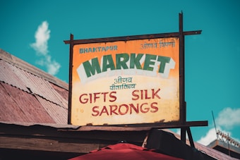 A vintage and rustic signboard with the text 'Bhaktapur Market', advertising items such as gifts, silk, and sarongs. The sign has a distressed look, mounted on a wooden structure. The background features a sky with fluffy clouds, and wooden rooftops.