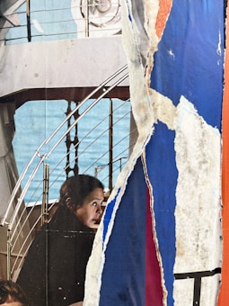 A collage combining elements of a torn poster featuring a woman gazing to the side, partially obstructed by layers of ripped paper. The background includes railing and industrial structures, giving an urban feel.