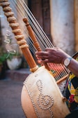 A close-up of a musical instrument featuring wooden pegs and strings, held by a person with a watch on their wrist. The body of the instrument is decorated with intricate patterns resembling musical symbols, crafted with beads. The setting appears to be an outdoor area with a blurred background.