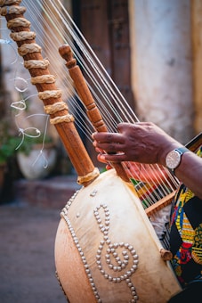 A close-up of a musical instrument featuring wooden pegs and strings, held by a person with a watch on their wrist. The body of the instrument is decorated with intricate patterns resembling musical symbols, crafted with beads. The setting appears to be an outdoor area with a blurred background.