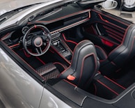 A luxurious car interior featuring black leather seats with red stitching and accents. The dashboard has a sleek, modern design with a digital display, and the steering wheel is sporty with a prominent logo. The center console includes futuristic controls and gear selector, emphasizing a high-end, sophisticated look.