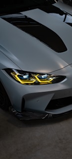 A sleek and modern car front with a metallic finish. The headlights are illuminated with a striking yellow hue, and the hood features a prominent black scoop. The car appears to be parked indoors on a dark concrete floor.