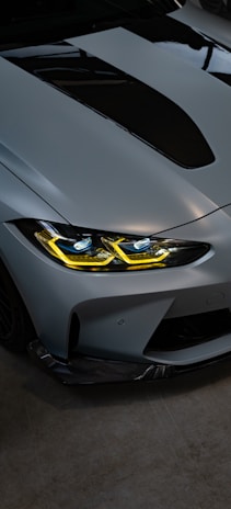 A sleek and modern car front with a metallic finish. The headlights are illuminated with a striking yellow hue, and the hood features a prominent black scoop. The car appears to be parked indoors on a dark concrete floor.