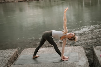 A person practices yoga on a mat by a calm river, performing a reverse triangle pose with one arm extended upwards and the other touching the ground. The setting is serene with a focus on balance and flexibility.