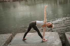 A person practices yoga on a mat by a calm river, performing a reverse triangle pose with one arm extended upwards and the other touching the ground. The setting is serene with a focus on balance and flexibility.