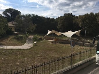 A grassy area with a small pathway leading to a structure with beige tensile canopy shelters. The surrounding environment is lush with dense trees and a small building with a tiled roof on the left. There is a black wrought iron fence in the foreground and part of a vehicle is visible on the right.
