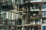 Reflections of various apartment balconies and windows are seen in the glass facade of a modern building, creating a distorted and abstract pattern. The reflective surface captures multiple layers of architectural elements, blending them into an intricate mosaic.