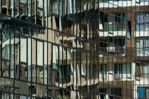 Reflections of various apartment balconies and windows are seen in the glass facade of a modern building, creating a distorted and abstract pattern. The reflective surface captures multiple layers of architectural elements, blending them into an intricate mosaic.
