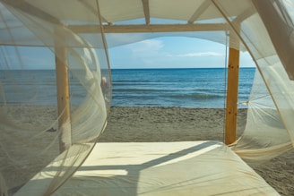 A peaceful beach scene viewed through sheer, flowing white curtains on a wooden canopy bed. The gentle waves of the ocean meet the sandy shore under a clear blue sky, creating a tranquil atmosphere.