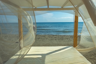 A peaceful beach scene viewed through sheer, flowing white curtains on a wooden canopy bed. The gentle waves of the ocean meet the sandy shore under a clear blue sky, creating a tranquil atmosphere.