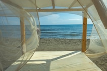 A peaceful beach scene viewed through sheer, flowing white curtains on a wooden canopy bed. The gentle waves of the ocean meet the sandy shore under a clear blue sky, creating a tranquil atmosphere.