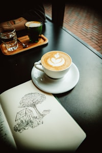A cappuccino with latte art sits on a white saucer next to an open sketchbook displaying a detailed drawing of a tree and a small house. Nearby, a glass of water and a small glass of espresso rest on a wooden tray, set on a dark table by a window overlooking a brick pathway.