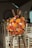 A colorful bouquet of flowers arranged on a wooden stand in an indoor setting with white chairs in the background. The bouquet features an assortment of flowers, primarily in shades of orange, red, and white with yellow accents.