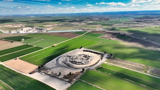 Aerial view of expansive farmlands featuring a prominent circular archaeological site. The fields are divided into various shades of green and brown, contrasted by the complex central structure with visible pathways and parking area.