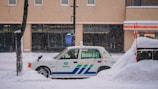 A taxi is parked along a snowy street with a thick layer of snow surrounding it. The background features a brick building with large windows, and snow is gently falling, creating a serene winter scene.